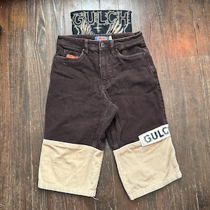 May include: Dark brown corduroy pants featuring a color-blocked design with tan lower legs. The pants include a button closure, a small red logo patch on the front pocket, and a white rectangular patch on the right leg with the text GULCH.