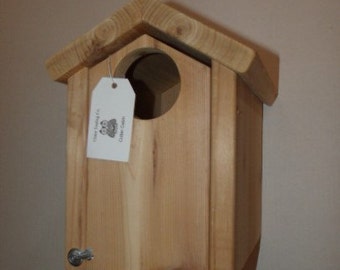 May include: A wooden birdhouse with a small round opening for birds to enter. The birdhouse is painted a natural wood color and has a small metal latch on the bottom. The birdhouse is labeled "Timber Trading Co. Cedar Cabin".