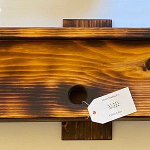 May include: A wooden birdhouse with a burnt finish and a small hole for birds to enter. The birdhouse has a label that reads "Urban Trading Co. Cypress Cabin".