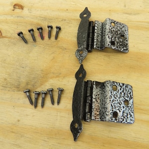 May include: Two decorative black metal hinges with textured silver-colored plates and matching screws are arranged on a light wood surface. The hinges have a decorative, ornate design and are likely for a cabinet or door.