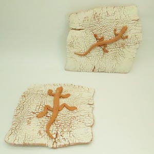 May include: Two ceramic tiles with a textured, off-white surface and a brown lizard design. The lizards are in a crawling position. The tiles have a rustic, handmade appearance, and are likely decorative wall art.