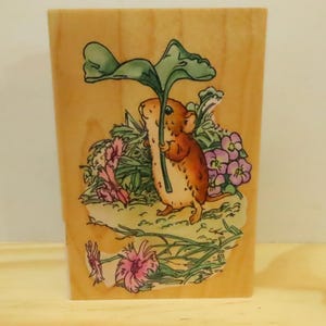 May include: A wooden block with a charming illustration of a brown mouse sheltering under a large green leaf. The mouse is amidst pink and purple flowers and green foliage, set against a light wood grain background.