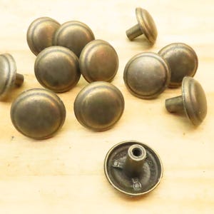 May include: A collection of eleven round, brass-toned metal furniture knobs. Each knob features a smooth, domed top surface with a slightly weathered, antique finish. One knob is positioned upside down to reveal the central threaded mounting post.
