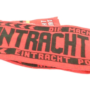 May include: Red and black knitted scarf with the word "Eintracht" in large black letters. The scarf also features the words "Die Macht" and "Eintracht Pow" in black. The scarf is folded and lies on a white surface.