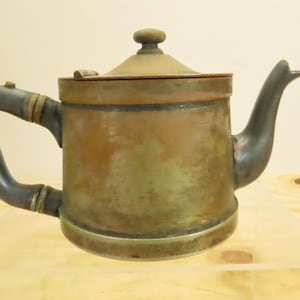May include: Antique copper teapot with a dark handle and spout. The teapot has a round body, a lid with a small knob, and brass accents. The handle is curved, and the spout is angled upwards. The teapot is sitting on a light-colored surface.