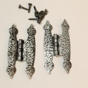 Pode incluir: Duas dobradiças decorativas de metal texturizado, com aspecto martelado, e um conjunto de parafusos. As dobradiças são prateadas com um design ornamentado e pontiagudo. Os parafusos são cinza escuro.
