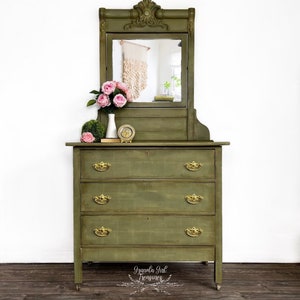 May include: A green antique dresser with a mirror and three drawers. The dresser has gold hardware and is sitting on a wooden floor. The dresser is decorated with a vase of pink flowers and a small clock.