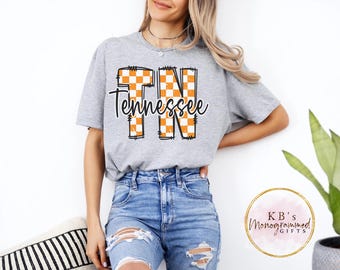 May include: A gray t-shirt with the word "Tennessee" in a black script font. The letters "TN" are filled with an orange and white checkered pattern. The shirt is worn with blue jeans.