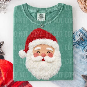 May include: A sage green t-shirt featuring an embroidered Santa Claus face with a red hat and white beard. The shirt has a crew neck and short sleeves. A silver necklace with a sun pendant is visible.