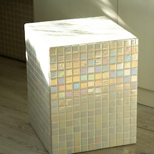 May include: A square, iridescent mosaic tile cube. The cube is made of small, square tiles that reflect light, creating a shimmering effect. The cube is a decorative accent piece, possibly a stool or side table.