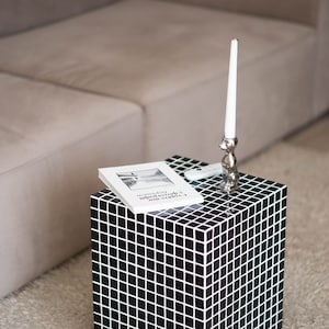 May include: A black and white checkered cube-shaped side table with a book and a silver candle holder on top. The table is sitting on a light brown rug.