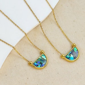 May include: Three gold necklaces with a half-circle pendant made of iridescent abalone shell. The necklaces are on a white surface.