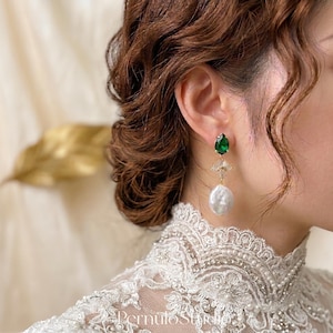 Puede incluir: Pendientes colgantes elegantes con una gema verde en forma de pera, un delicado detalle floral dorado y una gran perla blanca irregular. Llevados por una persona con cabello castaño y un top de encaje blanco.