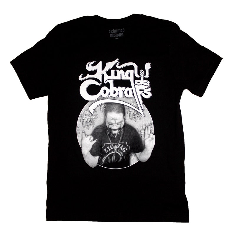 May include: Black t-shirt with a white graphic of a man with the text "King Cobras" above him. The text "exhumed designs" is printed in white on the top of the shirt.