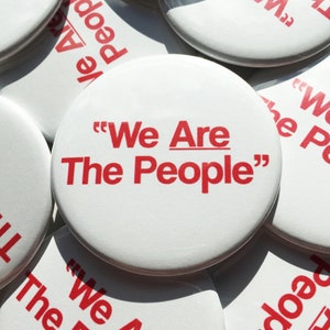 May include: A collection of white round buttons with red text that reads "We Are The People" in quotation marks.