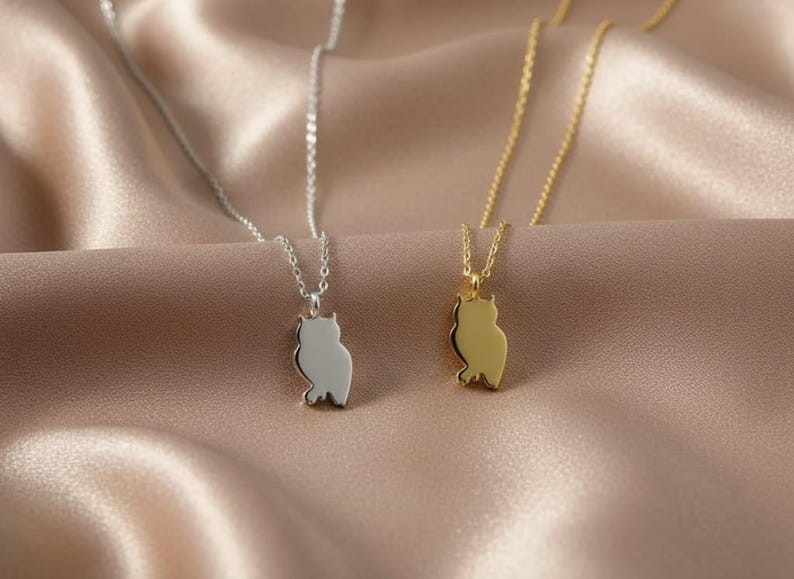 May include: Two owl pendant necklaces, one silver-toned and one gold-toned, rest on a satin surface. The pendants are shaped like owls. The necklaces are delicate and have a simple design, suitable for everyday wear.