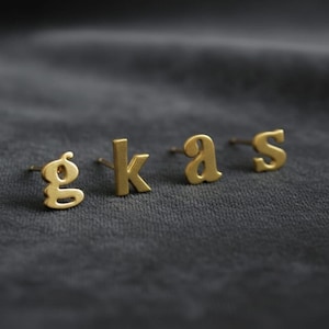 May include: Four small, gold-colored letter stud earrings spelling out the word "gkas". The letters are in a simple, sans-serif font and are displayed on a dark gray, textured surface. The earrings are evenly spaced.