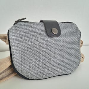 Può includere: Un piccolo portafoglio in tessuto grigio intrecciato con cerniera nera e chiusura a scatto in pelle nera. Il portafoglio ha una forma arrotondata e un bordo nero. Sfondo bianco con un pezzo di legno alla deriva.