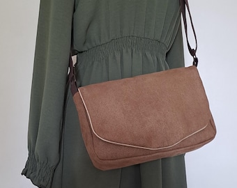 Op de afbeelding: Een bruine messenger bag van suède-achtig materiaal met een klepsluiting en een bruine schouderriem. De tas heeft een lichtgekleurde rand en wordt getoond op een mannequin die een olijfgroene jurk met lange mouwen draagt.