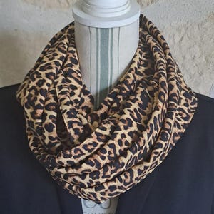 Pode incluir: Cachecol infinito com estampa de leopardo, com manchas marrons em fundo bege. O cachecol é drapeado ao redor do pescoço de um manequim, mostrando seu design circular e tecido macio. Um acessório de moda versátil.