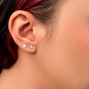 May include: A pair of silver stud earrings, one shaped like a star and the other a crescent moon, are displayed on an ear. The earrings are small and minimalist, offering a subtle celestial accent. The ear is set against a blurred background.