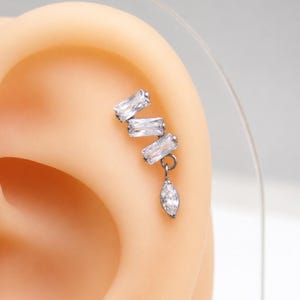 May include: A silver-toned ear cartilage piercing featuring three clear baguette-cut stones arranged in a stepped, diagonal pattern, with a single dangling marquise-cut clear stone suspended from the bottom of the lowest baguette stone.