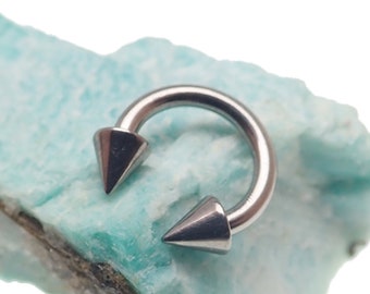 May include: A silver-toned circular barbell body jewellery piece featuring two pointed cone-shaped ends. The metal ring has a smooth, polished finish and is shaped into a horseshoe curve, with the sharp conical spikes facing inward toward each other.