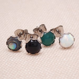 May include: Four stud earrings featuring round, scalloped-edge metal settings. The set includes one iridescent dark gray stone, one solid black stone, one deep green stone, and one white stone with subtle iridescent flecks, all with metal backs.