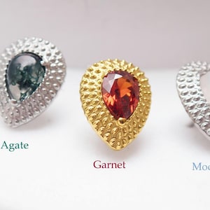 Pode incluir: Close-up de três brincos de pedra preciosa. Os brincos apresentam pedras em forma de pêra em ágata musgo, granada e pedra da lua, cada uma emoldurada em uma estrutura de metal texturizada. As pedras preciosas são rotuladas com seus nomes.