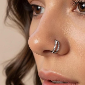 May include: A close-up of a person's nose with a double-hoop nose ring. The nose ring is silver-toned and features two rows of small, clear gemstones. The skin around the nose is smooth and has a natural tone.
