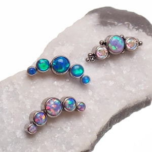 May include: Three sets of opal and crystal stud earrings. Each set features a curved design with multiple round stones in shades of blue, green, and pink. The earrings are set on a silver-colored metal.