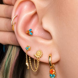 May include: A collection of gold-tone ear jewellery including a multi-coloured crystal stud, a shell-shaped stud with two attached chains, a small hoop with a dangling crystal charm, and two thin hoops featuring clear and orange crystals.