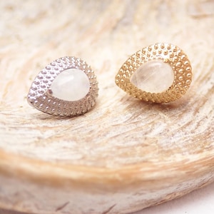 May include: A pair of teardrop-shaped stud earrings. One is silver-coloured, the other gold-coloured, both with a textured, dotted border. Each earring has a smooth, oval-shaped, milky white stone centre. The earrings are on a light-coloured, textured surface.