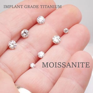 May include: A hand displays several round, clear moissanite gemstones of varying sizes. The text "IMPLANT GRADE TITANIUM" and "MOISSANITE" are visible. The gemstones are set against a light background.