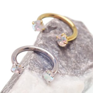 May include: Two horseshoe-shaped body jewellery pieces. One is gold-toned, the other silver-toned. Each has a clear, iridescent gemstone at each end. The jewellery is displayed on a grey, textured stone.