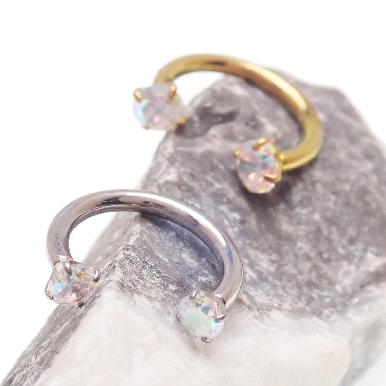 May include: Two horseshoe-shaped body jewellery pieces. One is gold-toned, the other silver-toned. Each has a clear, iridescent gemstone at each end. The jewellery is displayed on a grey, textured stone.