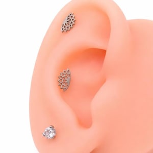 May include: Close-up of an ear with three pieces of jewelry. Two are silver-colored, half-circle earrings with a floral design. The third is a small, silver-colored stud with a clear, round gemstone. The ear is a light peach color.