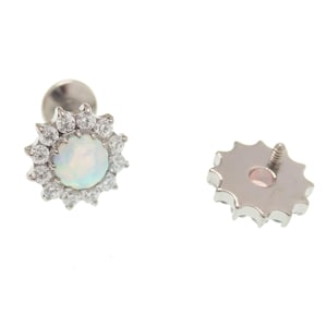 Puede incluir: Una pieza de joyería corporal de plata con una piedra de ópalo blanco rodeada de piedras de circonita cúbica transparentes. La parte posterior de la pieza de joyería es un diseño de estrella de plata.