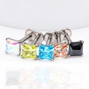 May include: Five labret studs with square-cut gemstones in iridescent white, lime green, bright blue, peach, and solid black. Each piece features a metallic post and a flat circular backing, arranged in a row to display the varied stone colors.