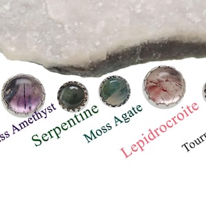 Könnte beinhalten: Eine Sammlung von fünf Edelstein-Cabochons, darunter Stained Glass Amethyst, Serpentine, Moosachat, Lepidocroite und Turmalinquarz, jeweils in einer silberfarbenen Lünette gefasst. Die Edelsteine werden vor weißem Hintergrund präsentiert.