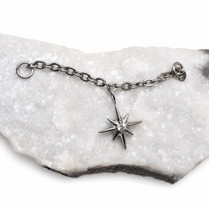 May include: A silver-toned metal chain featuring a dangling star-shaped charm. The charm has eight pointed rays and a small, clear, round gemstone set in the center. The chain includes a circular jump ring at each end for attachment purposes.