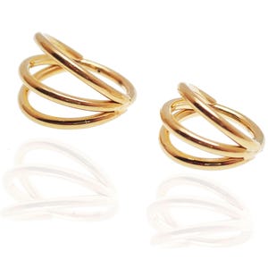 May include: Two gold-colored ear cuffs, each featuring a triple-band design. The bands are smooth and rounded, creating a modern, minimalist aesthetic. The ear cuffs are displayed on a white surface, with their reflections visible below.