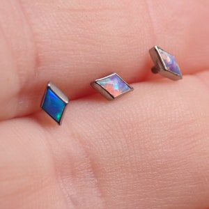 May include: Three diamond-shaped opal body jewellery pieces in different colours. The pieces are made of titanium and have a silver finish. The opals are blue, pink, and white.