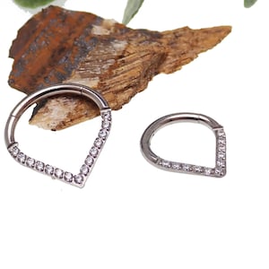 May include: Two silver teardrop-shaped body jewelry pieces with a row of clear gemstones around the edge.
