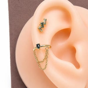 May include: A gold ear cuff featuring a teardrop-shaped dark stone and two delicate hanging chains, paired with a separate gold stud earring set with three small, rectangular, light green stones arranged in a cluster on the upper ear cartilage.