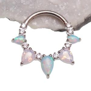 Pode incluir: Um piercing de septo prateado com uma fileira de pequenas pedras preciosas transparentes e pedras opalescentes em forma de pêra. As pedras opalescentes têm um brilho suave e iridescente. O piercing está sobre um fundo branco.