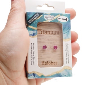 May include: A pair of pink titanium earrings in a small box. The box is white with blue and green accents. The box has the text "Halobes" and "Titanium" on it.