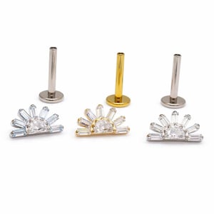 May include: Three pieces of jewelry: two silver-colored and one gold-colored. Each piece features a sunburst design with a central clear stone and surrounding baguette-cut stones. The posts are cylindrical. The jewelry is likely for body piercings.