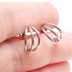 May include: A pair of silver-colored earrings. Each earring features a three-loop design, creating a layered effect. The earrings are small and appear to be made of a shiny metal. The design is minimalist and modern, suitable for everyday wear.