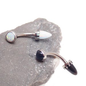 May include: Two curved, silver-toned body jewelry pieces. One features a white opal stone and a white, bullet-shaped end. The other has a black stone and a dark, bullet-shaped end. Both are on a gray stone surface.